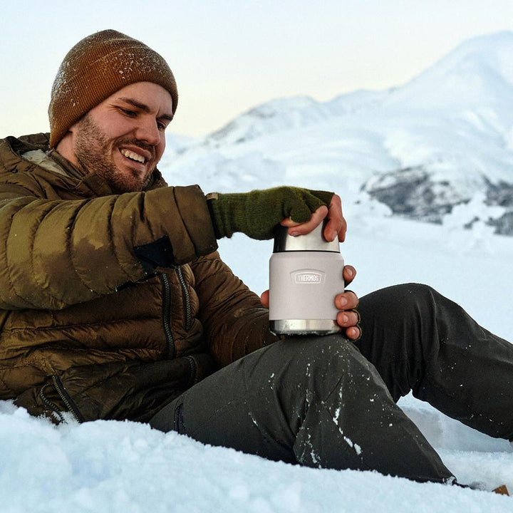 Man sitting on a snowy mountain, opening a Thermos 16oz Food Jar.