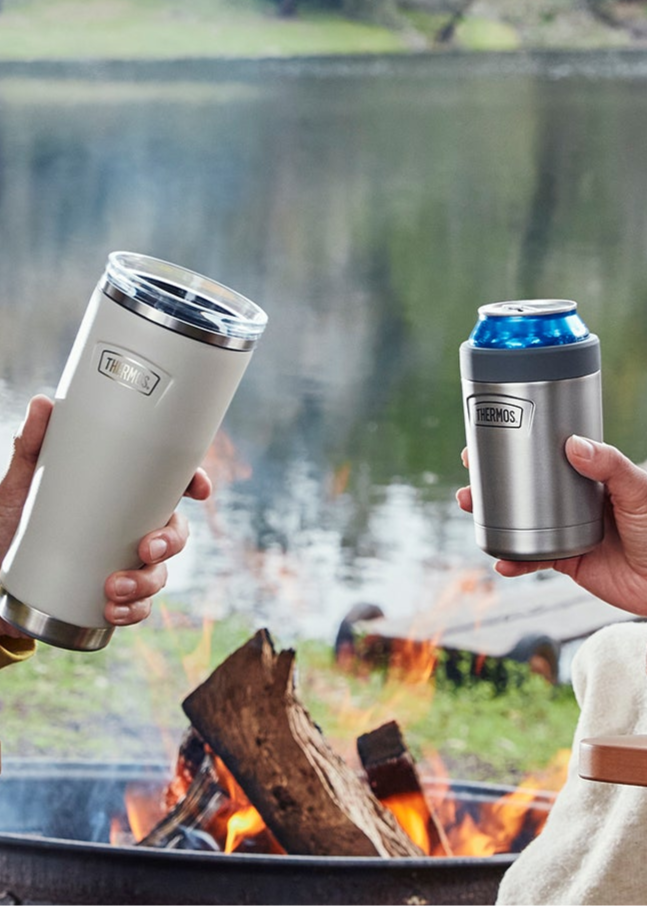 Two people holding insulated cups by a campfire with a lake and trees in the background