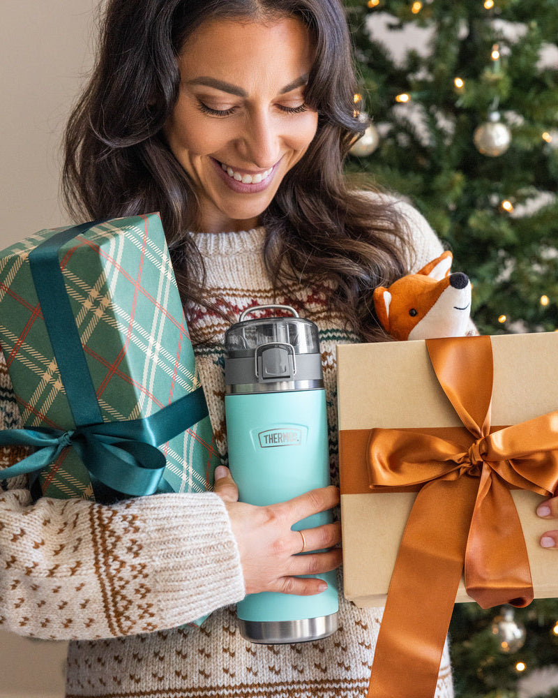 Woman holding a gift, thermos water bottle, and wrapped present in front of a Christmas tree.