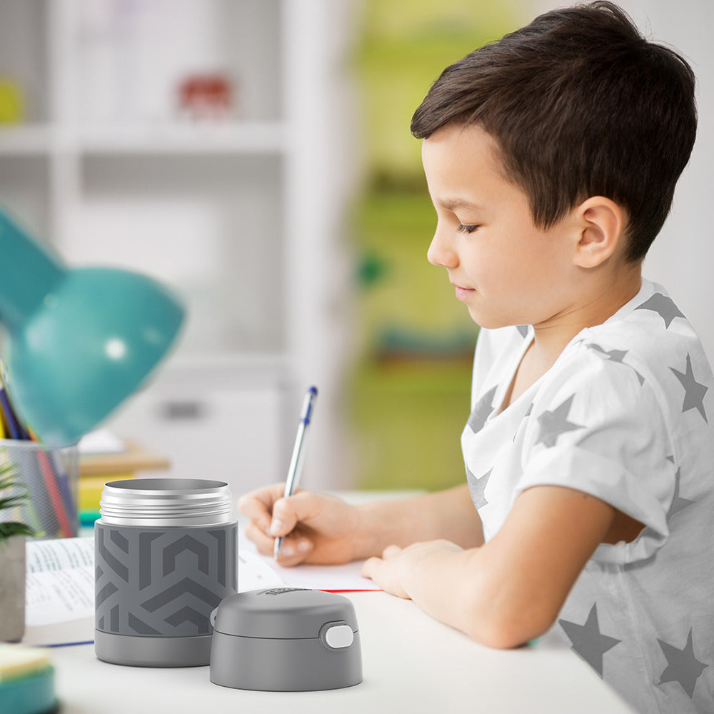 Boy sitting at table doing homework with an open 10 ounce Funtainer food jar in grey geometric pattern open next to him.