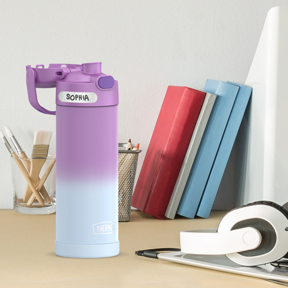 16-ounce Funtainer Stainless Steel Water Bottle with Spout, Purple Ombre, side view showing a sticker name label, lid open, sitting on a table.